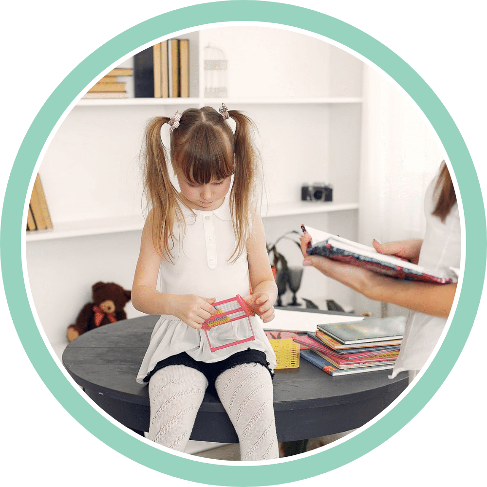 A young girl works on a learning activity at a table at home while her clinician reviews materials beside her